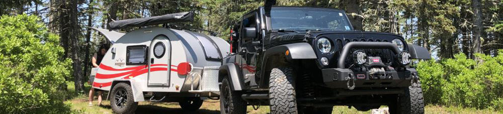 Black Jeep pulling red and white teardrop camper in a wooded area.