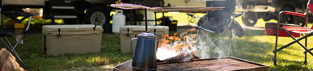 Campsite setup with stake grill over fire, coffee kettle, coolers in the background, and camping chairs.