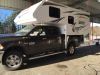 Black truck with white truck camper in the bed, parked in a parking lot.