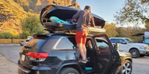 Person loading gear into a rooftop cargo box mounted on a black SUV at a scenic outdoor location.