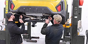 Two installers mounting a trailer hitch under a yellow vehicle on a lift.